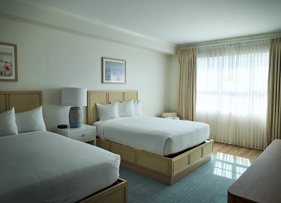 Two queen beds in a bright hotel room with a nightstand, lamps, artwork, and large windows with beige curtains.