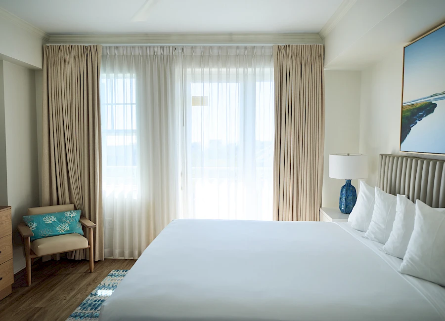 A bright hotel room with a large bed, white bedding, beige curtains, a nightstand lamp, a blue ceramic lamp, and a chair by the window.