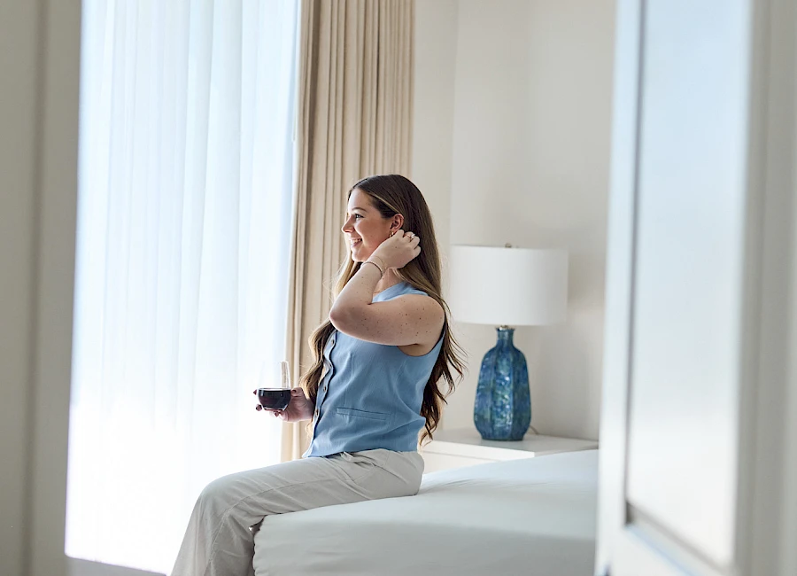 A woman sits on a bed in a bright hotel room, holding a phone and coffee, adjusting her hair by the door as light streams in.