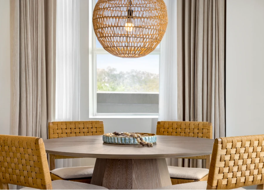 A round wooden dining table with four woven-back chairs in a bright room, a woven pendant light, and a window with sheer curtains.