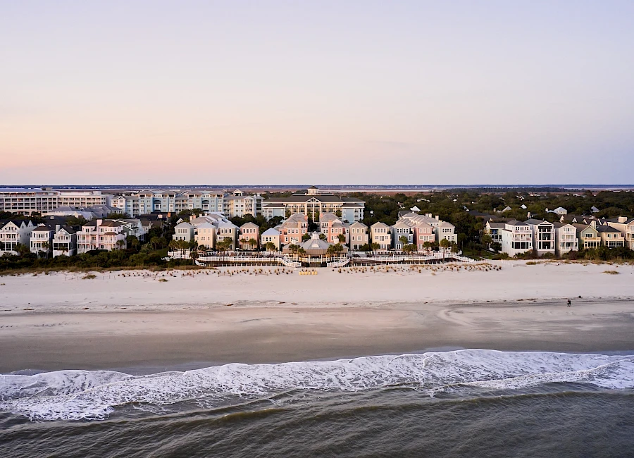 A row of pastel beach houses line a sandy shoreline with gentle waves and a calm, pastel-hued sky at sunset, forming a peaceful coastal scene.