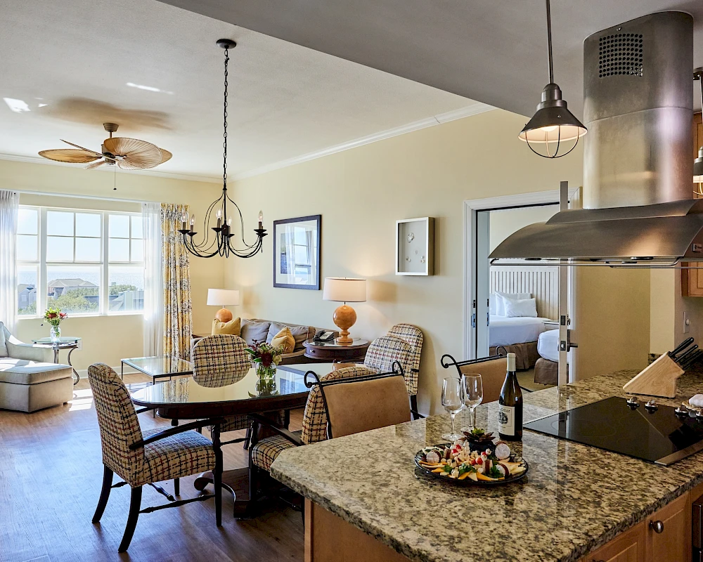 A cozy open-plan kitchen and dining area with a granite island, hanging pendant lights, a chandelier, a round dining table, and a bright sitting nook by a window.