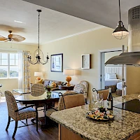A cozy open-plan kitchen and dining area with a granite island, hanging pendant lights, a chandelier, a round dining table, and a bright sitting nook by a window.