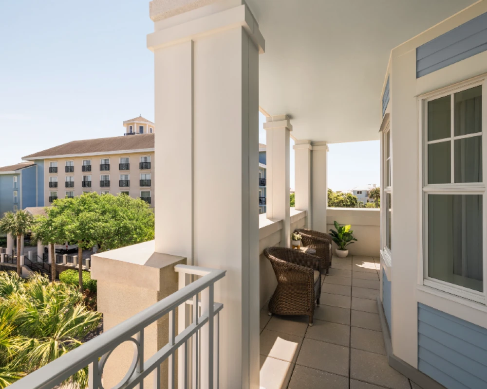 A bright seaside balcony with wicker chairs, pale blue siding, white columns, and views of palm trees and neighboring buildings in warm daylight.