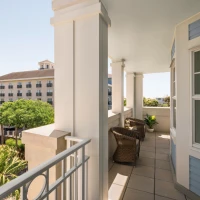 A bright seaside balcony with wicker chairs, pale blue siding, white columns, and views of palm trees and neighboring buildings in warm daylight.