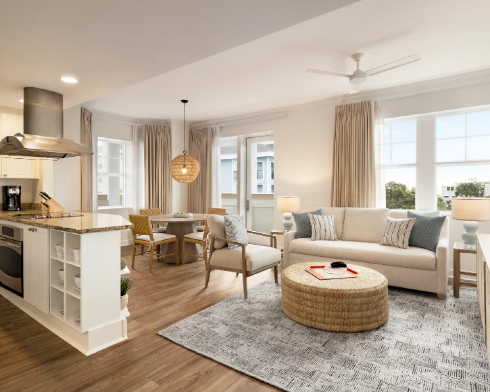 A bright, open-concept living room and kitchen with a cozy seating area, round wicker coffee table, neutral sofa, dining nook, and modern appliances—airy, stylish, and welcoming.