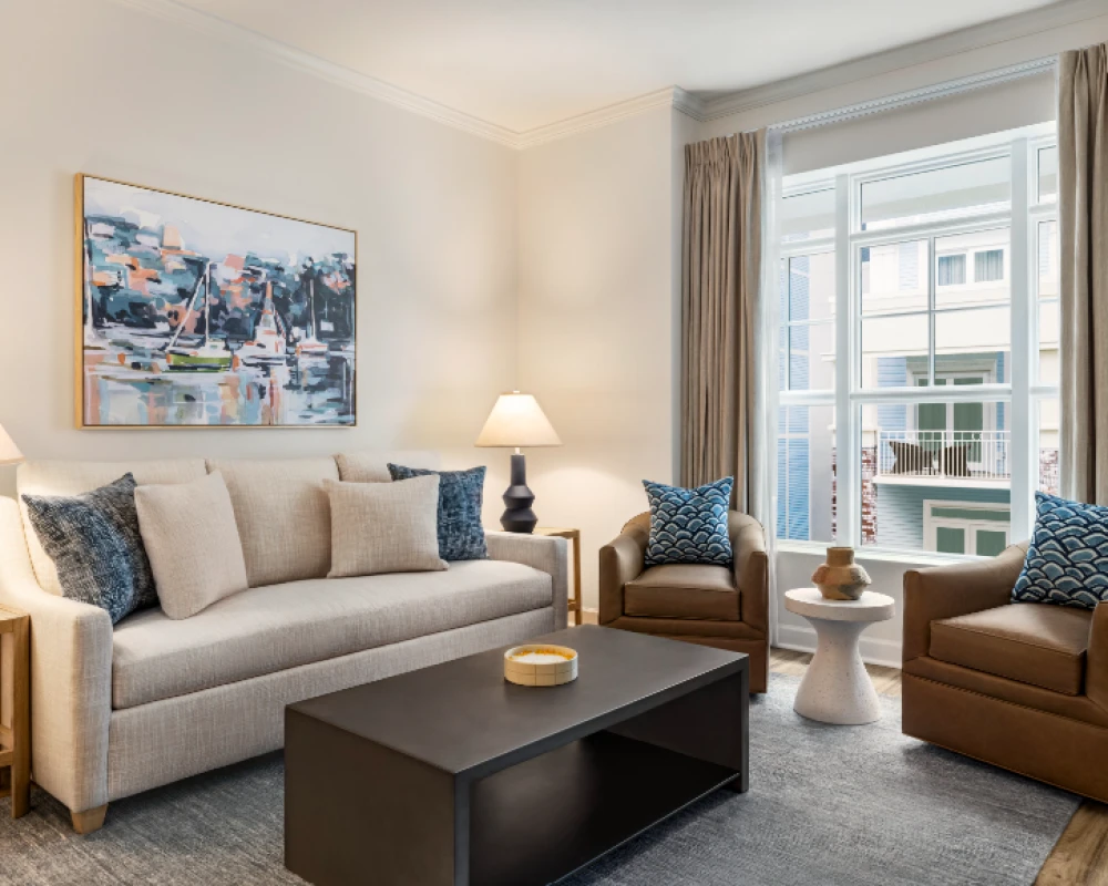 A cozy living room with a beige sofa, two brown armchairs, a dark coffee table, two table lamps, a painting on the wall, and a large window with beige curtains.