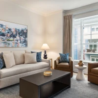 A cozy living room with a beige sofa, two brown armchairs, a dark coffee table, two table lamps, a painting on the wall, and a large window with beige curtains.