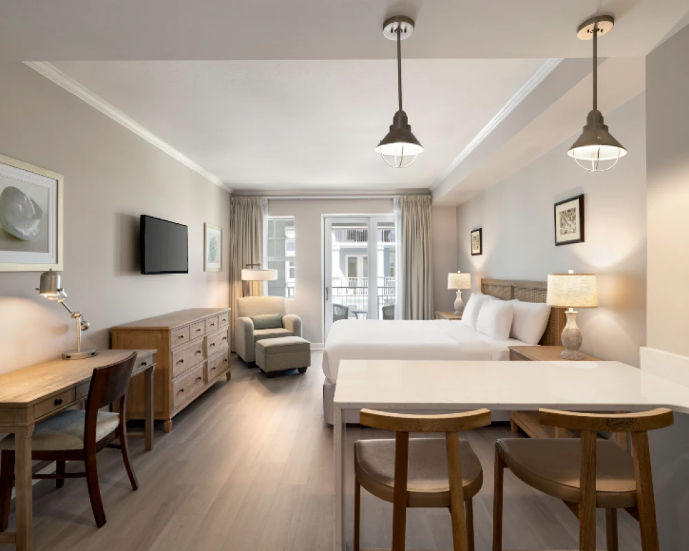 A modern hotel room with a king bed, bedside lamps, a seating area, wooden desk, wall art, a TV, and a small kitchenette with three stools by a breakfast bar.