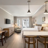 A modern hotel room with a king bed, bedside lamps, a seating area, wooden desk, wall art, a TV, and a small kitchenette with three stools by a breakfast bar.