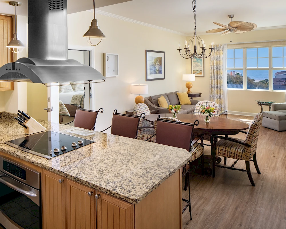 A modern open-concept kitchen and living room with a granite island, hanging pendant lights, stainless range, dining table, and a cozy seating area by large windows, all bathed in natural light.