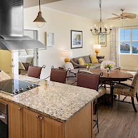 A modern open-concept kitchen and living room with a granite island, hanging pendant lights, stainless range, dining table, and a cozy seating area by large windows, all bathed in natural light.