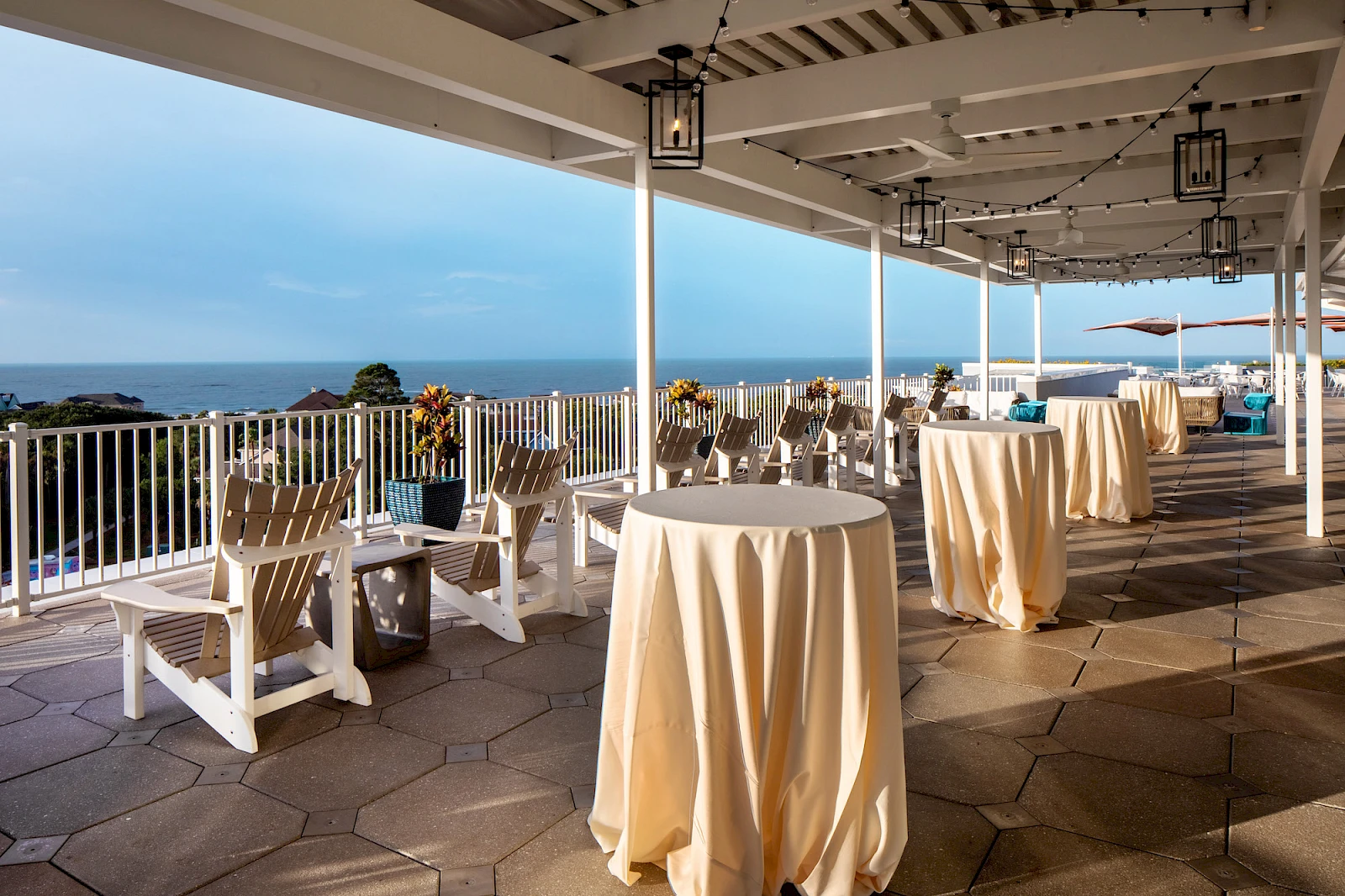 An oceanfront terrace with white patio furniture, high-top and low tables draped in cream cloths, and a sea view under a shaded pergola.