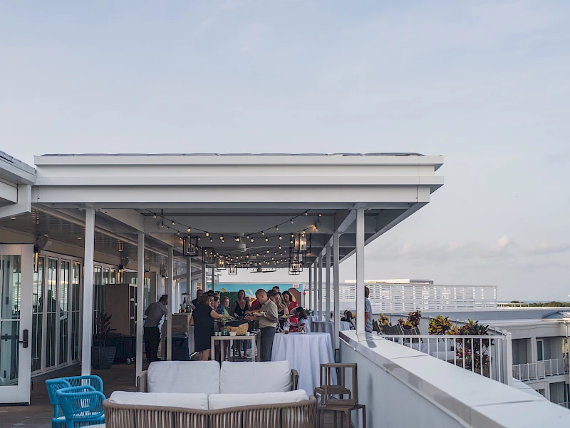 A rooftop party or lounge area with string lights, people mingling, white couches, and a view of the city skyline in the background, under clear skies.