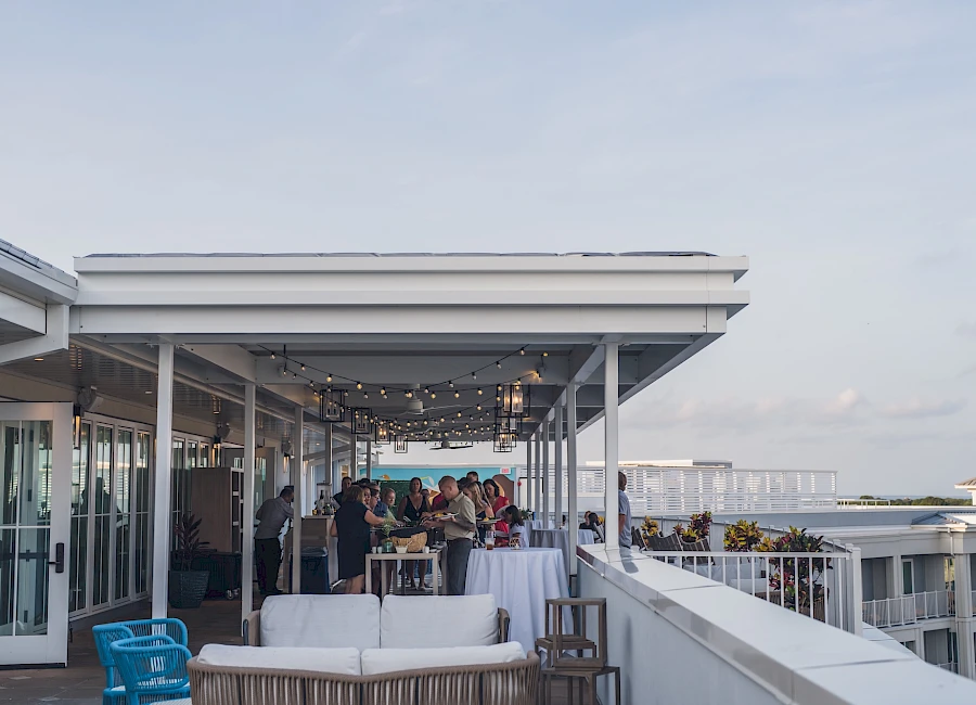 A rooftop party or lounge area with string lights, people mingling, white couches, and a view of the city skyline in the background, under clear skies.