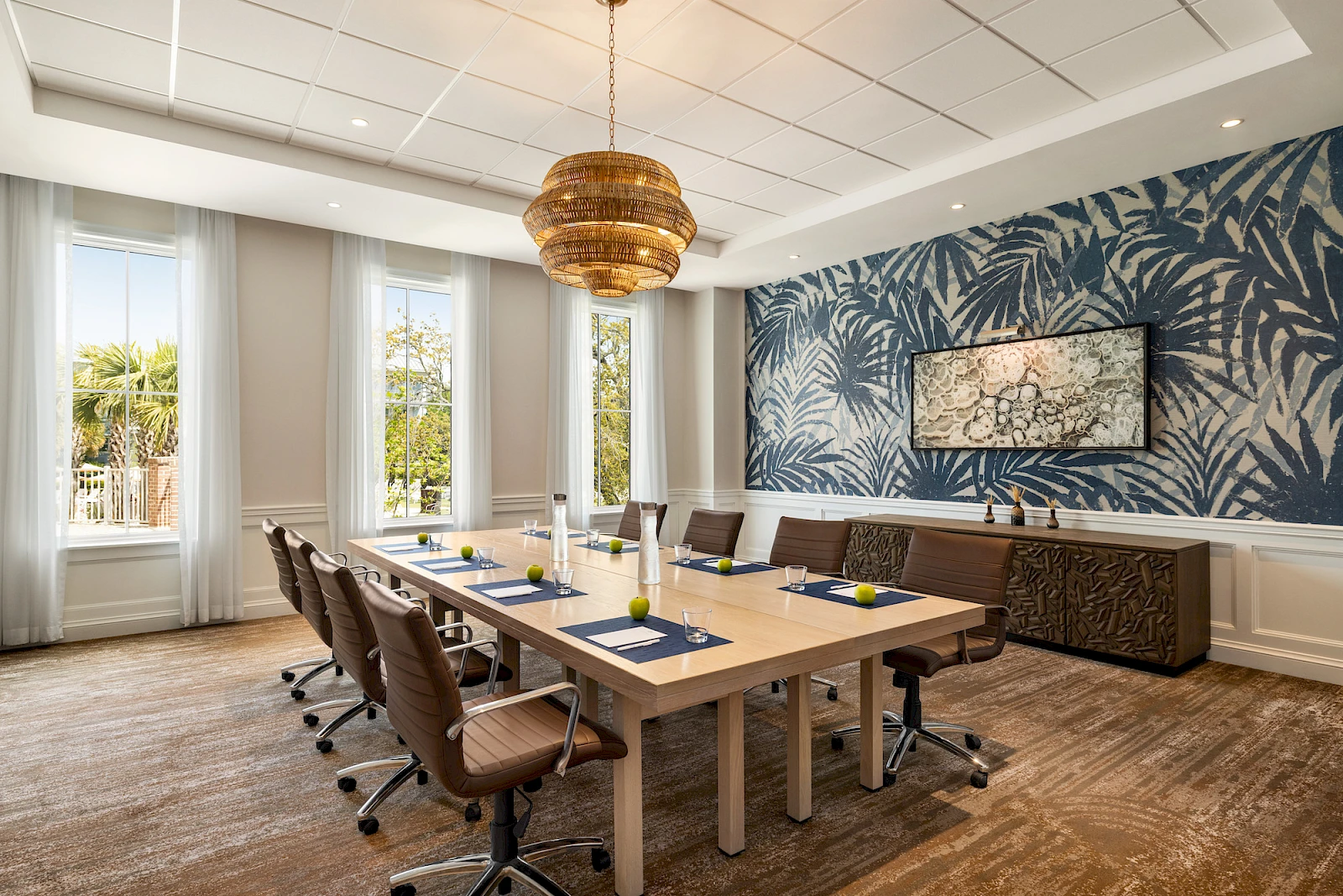 A modern conference room with a long wooden table, brown rolling chairs, tropical blue wallpaper, a hanging light, plants, and a side cabinet.