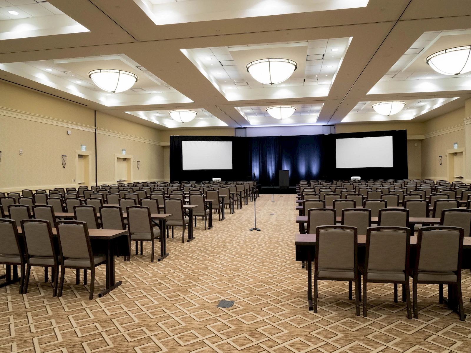 A large conference room set up for a presentation with rows of chairs facing a stage with two screens and blue lighting, ready for attendees.