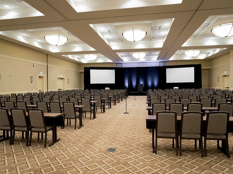 A large conference room set up for a presentation with rows of chairs facing a stage with two screens and blue lighting, ready for attendees.