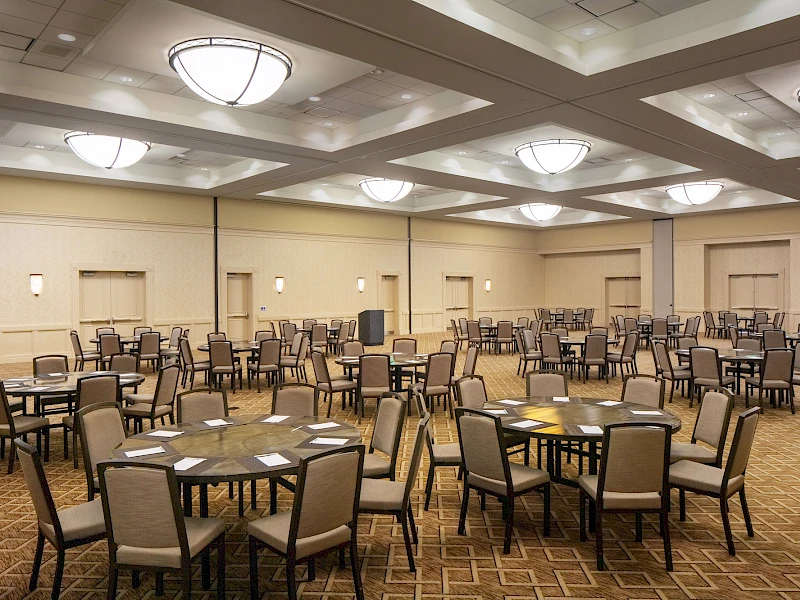 A spacious banquet hall set for a formal event with multiple round tables, chairs, and place settings, ready for dining and mingling.