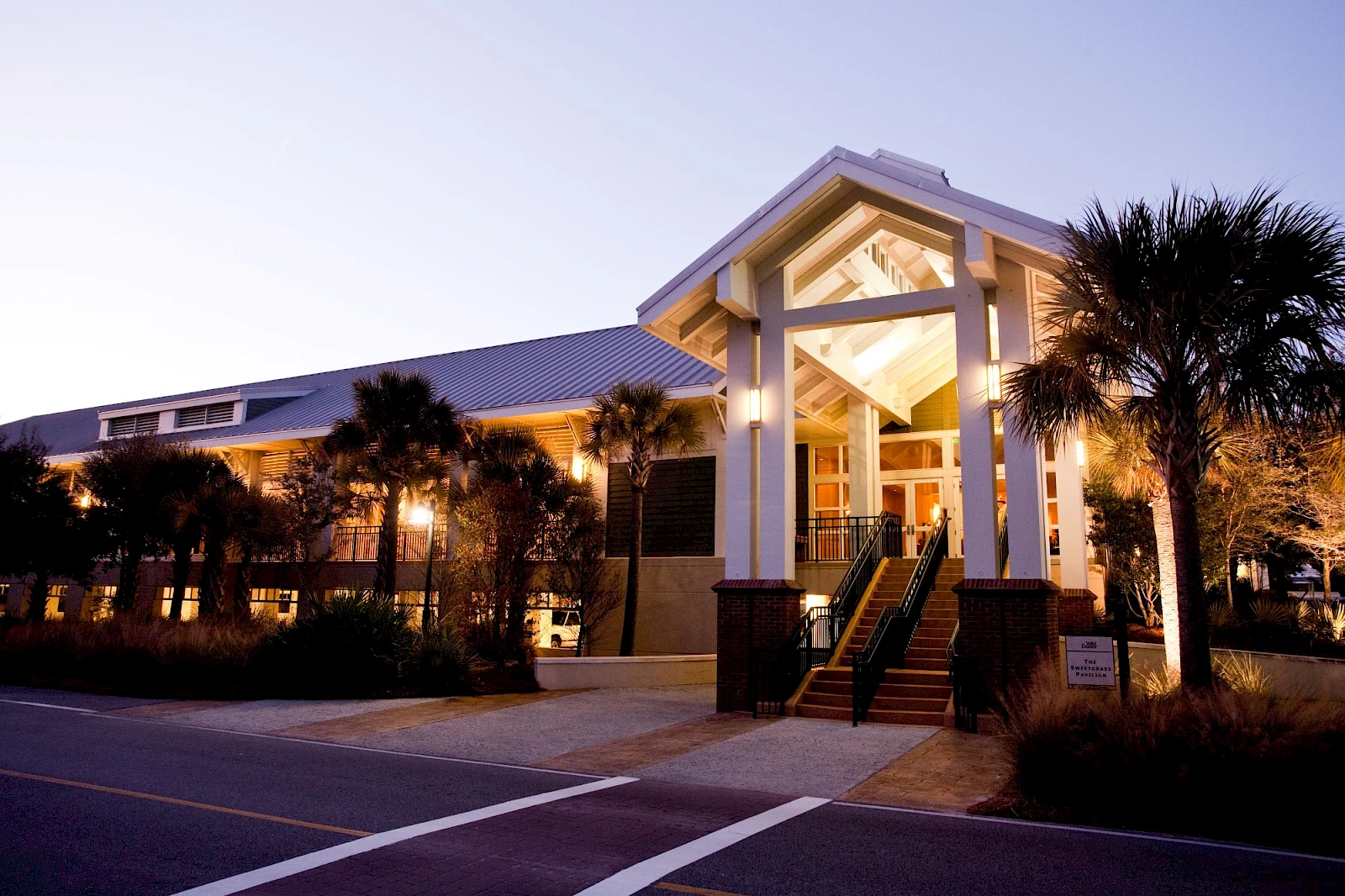 A sunny, modern seaside building with a tall portico, palm trees, and glass doors; outdoor lighting casts a warm glow at dusk.