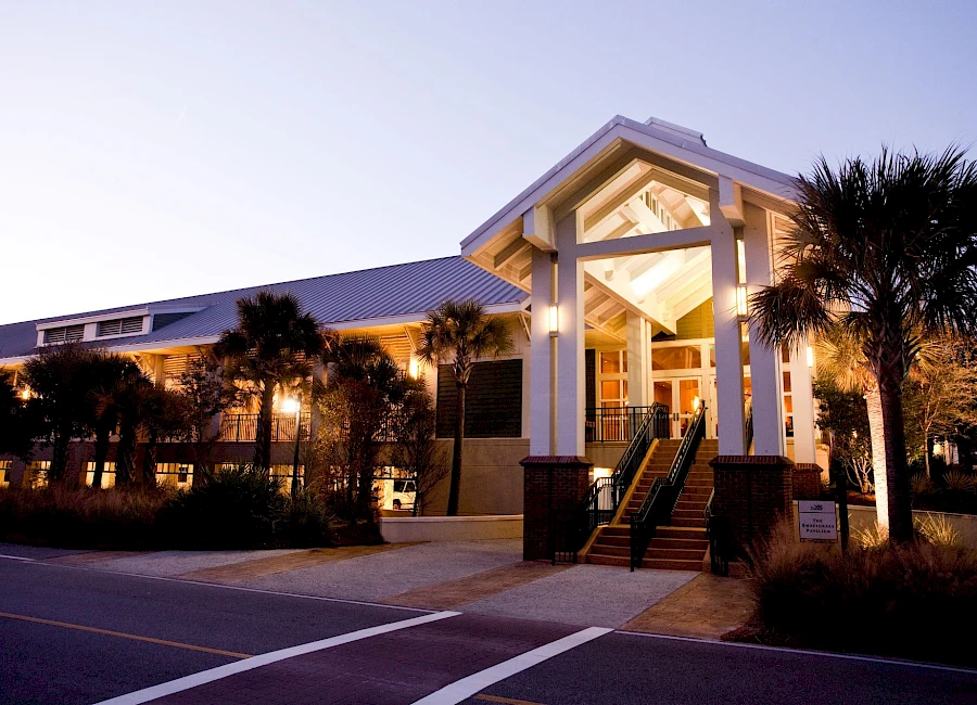 A sunny, modern seaside building with a tall portico, palm trees, and glass doors; outdoor lighting casts a warm glow at dusk.
