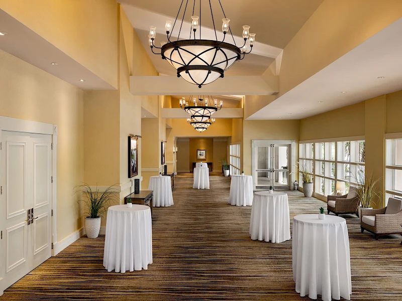 A spacious event hallway with beige walls, large windows, and round white-table cocktail setups; chandeliers hang above, creating a formal reception area.