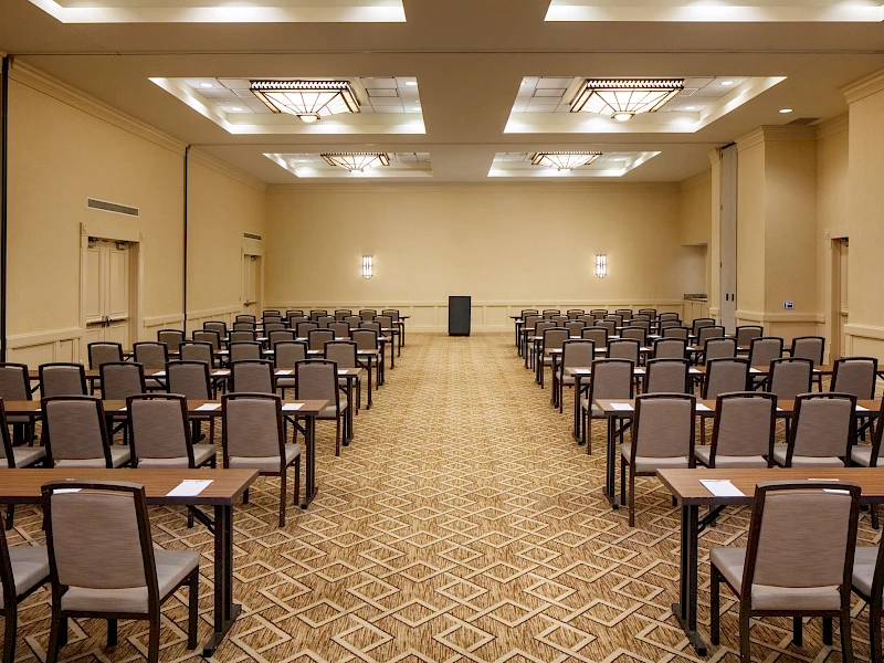 A large conference room set up with rows of chairs and desks facing a podium at the far end, under overhead lighting.