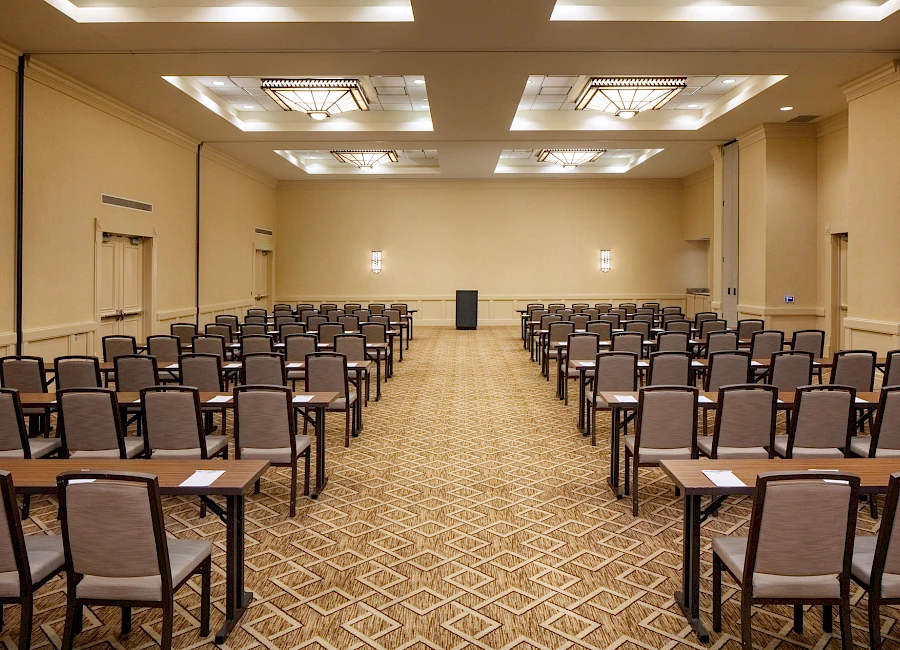 A large conference room set up with rows of chairs and desks facing a podium at the far end, under overhead lighting.