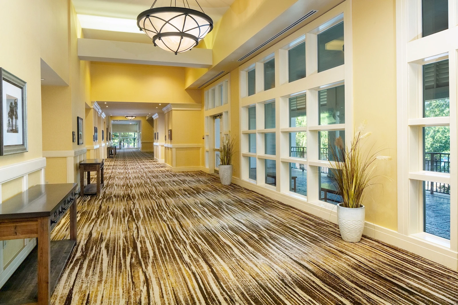 A bright hotel hallway with yellow walls, large windows, striped carpet, potted plants, and modern light fixtures, long and inviting.