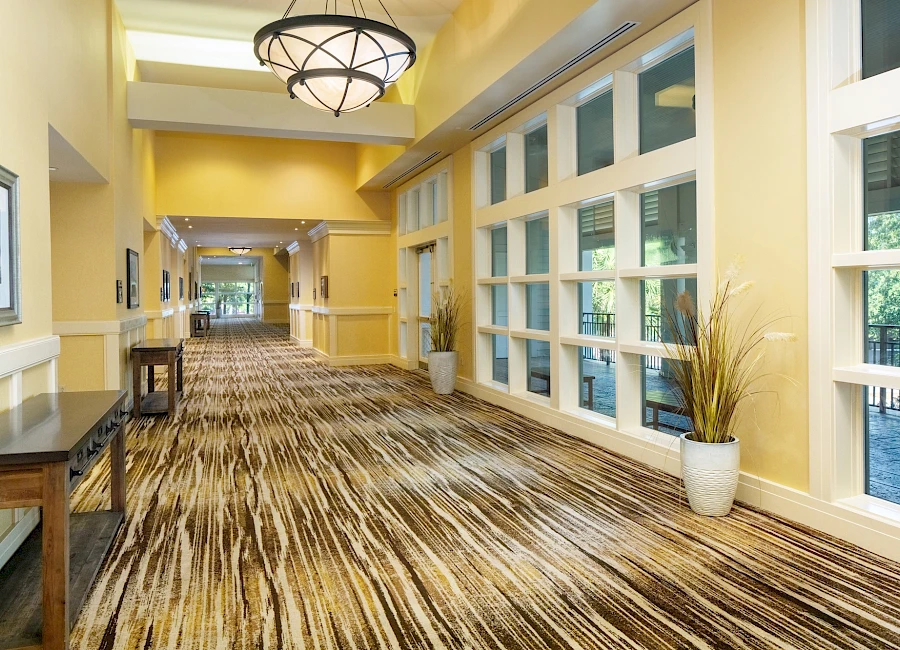 A bright hotel hallway with yellow walls, large windows, striped carpet, potted plants, and modern light fixtures, long and inviting.
