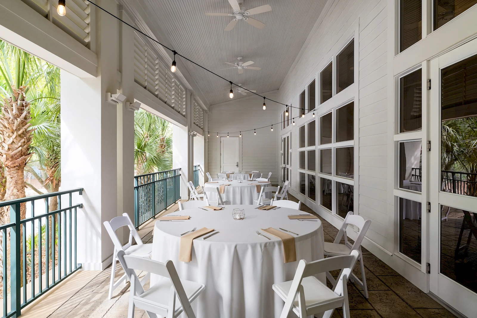 Alfresco dining area with white round tables, beige napkins, white folding chairs, string lights, and large windows on a covered patio overlooking greenery.