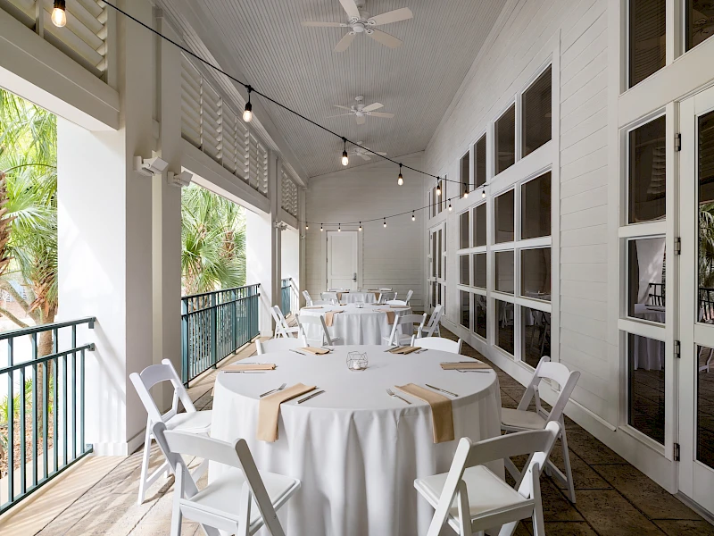 Alfresco dining area with white round tables, beige napkins, white folding chairs, string lights, and large windows on a covered patio overlooking greenery.