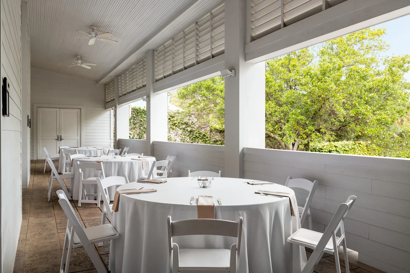 A bright, covered patio with white round tables and chairs, placed for outdoor dining, overlooking green trees through open windows.