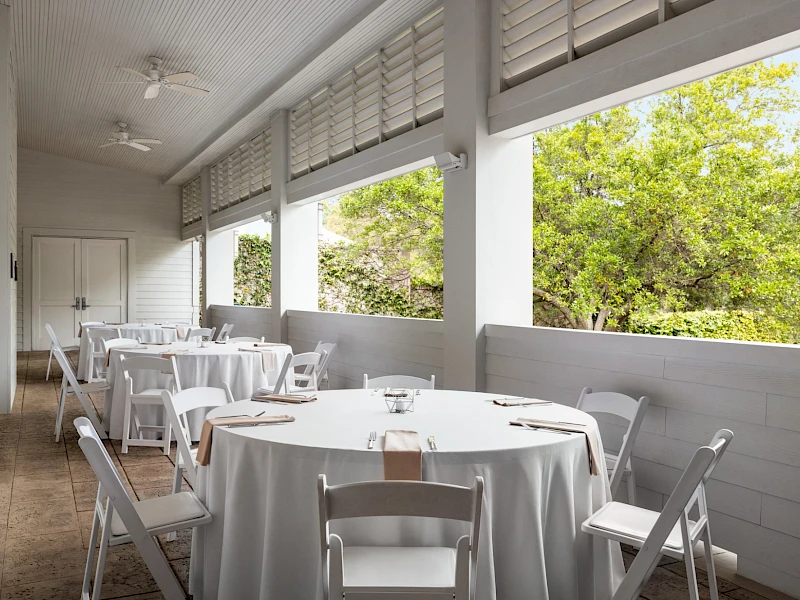 A bright, covered patio with white round tables and chairs, placed for outdoor dining, overlooking green trees through open windows.