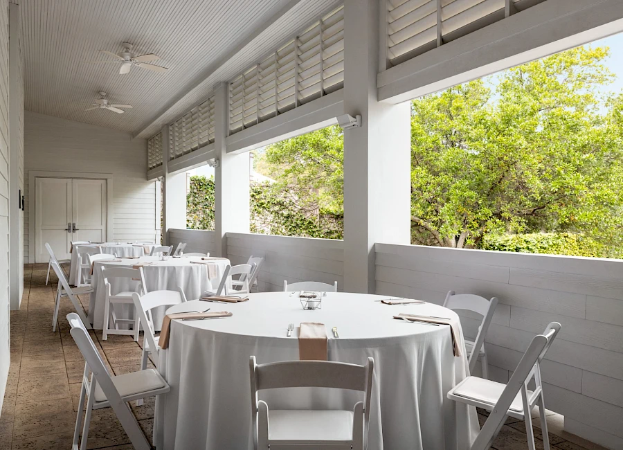 A bright, covered patio with white round tables and chairs, placed for outdoor dining, overlooking green trees through open windows.