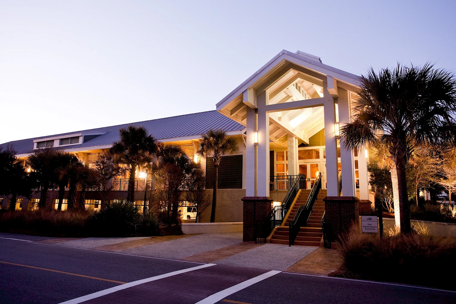 A bright, modern building with a welcoming entrance, white columns, tropical landscaping, and soft evening lighting along a paved street.