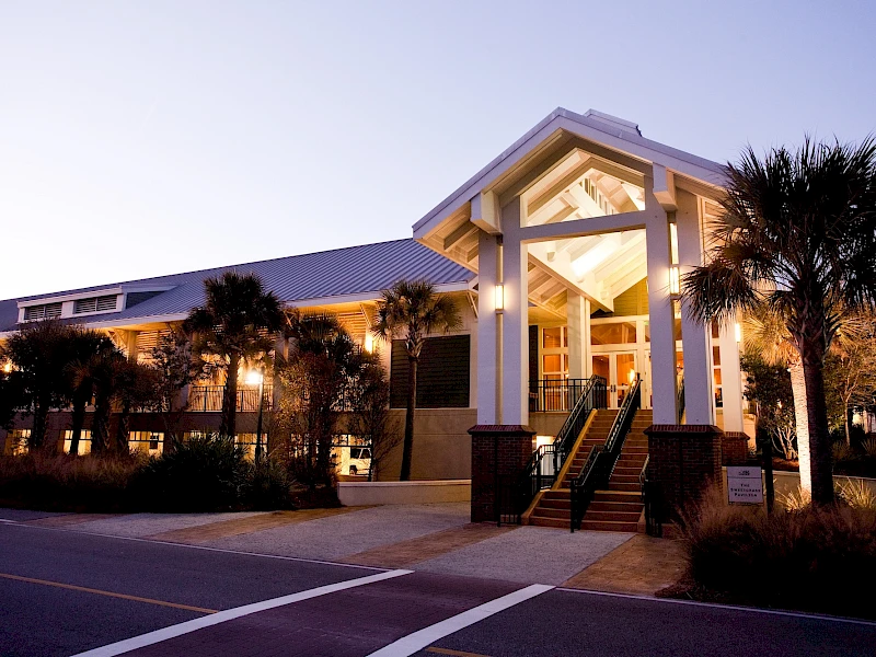 A bright, modern building with a welcoming entrance, white columns, tropical landscaping, and soft evening lighting along a paved street.