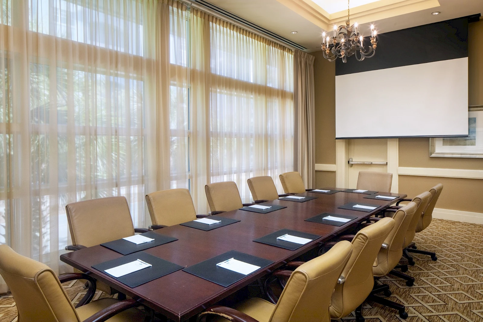 A formal conference room with a long table, beige chairs, large windows with sheer curtains, a projector screen, and a chandelier.