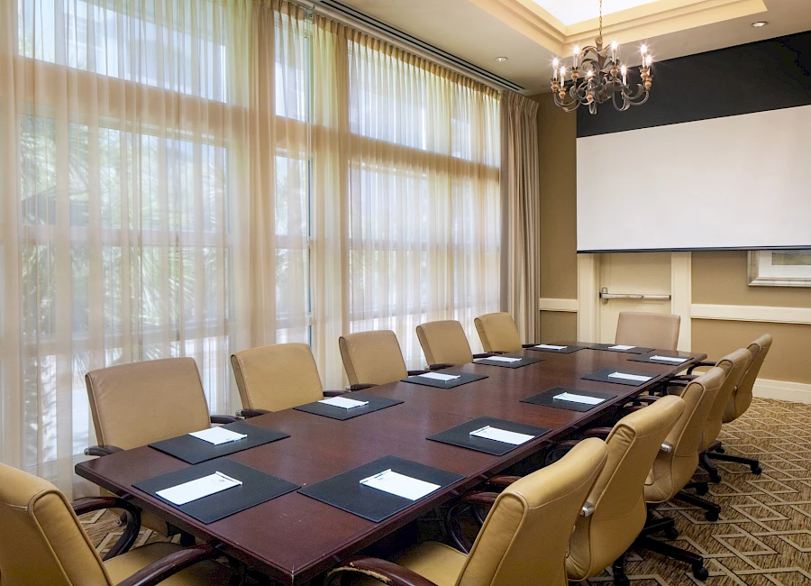 A formal conference room with a long table, beige chairs, large windows with sheer curtains, a projector screen, and a chandelier.