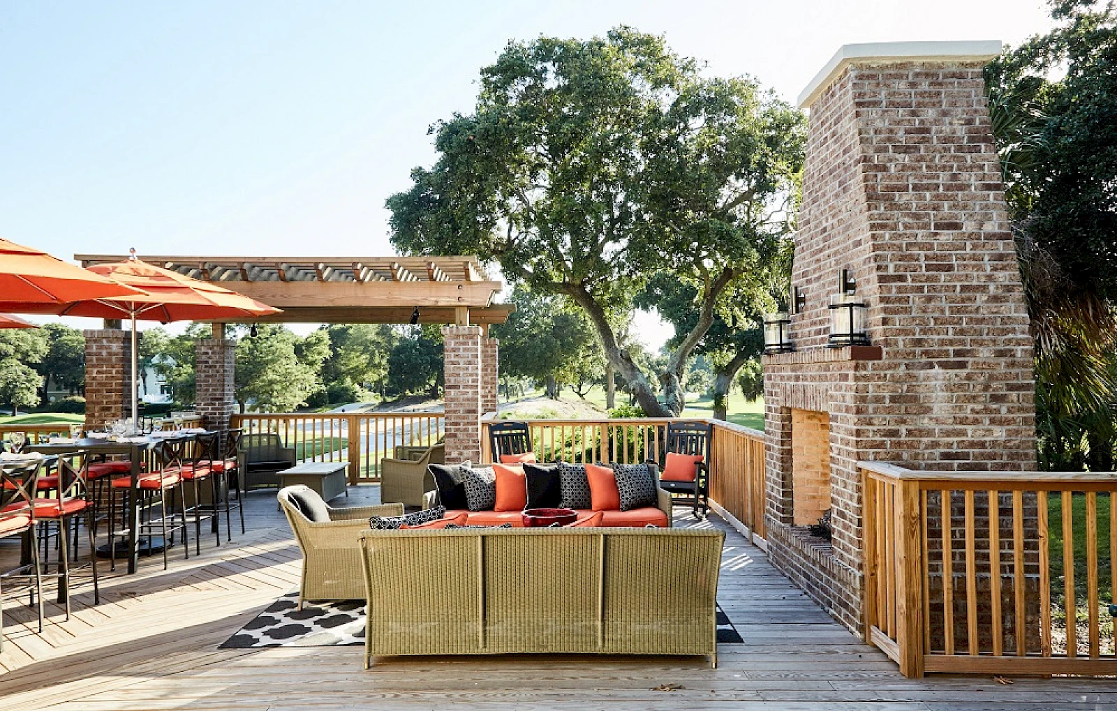 A sunny outdoor patio with wicker seating, orange umbrellas, a brick barbecue/oven, trees in the background, and a wooden railing.