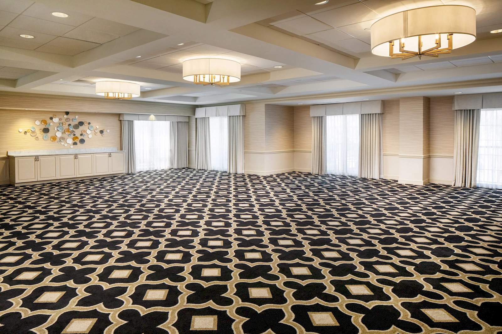 A large, empty conference/banquet room with patterned carpet, ceiling lights, draped windows, and a small side table along the far wall, ready for events.