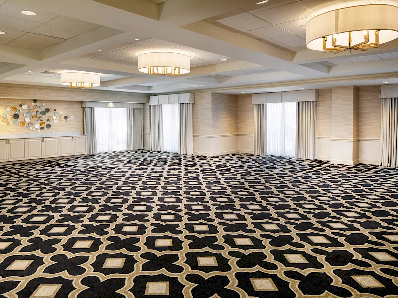 A large, empty conference/banquet room with patterned carpet, ceiling lights, draped windows, and a small side table along the far wall, ready for events.