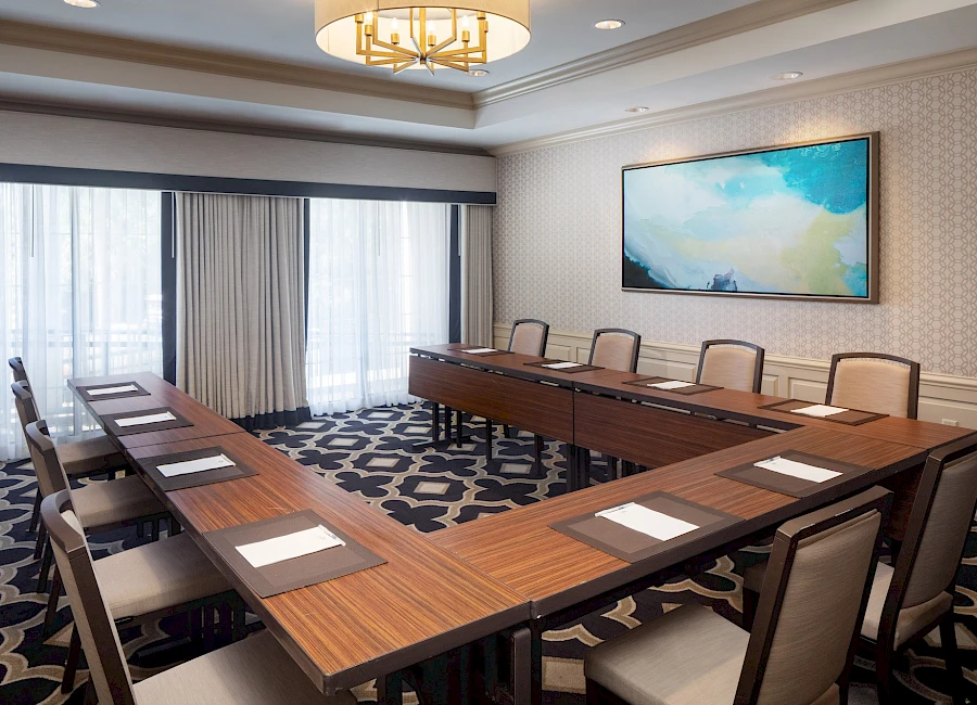 A modern conference room with a U-shaped wooden table, cushioned chairs, large windows with curtains, a patterned carpet, and a framed abstract painting on the wall.