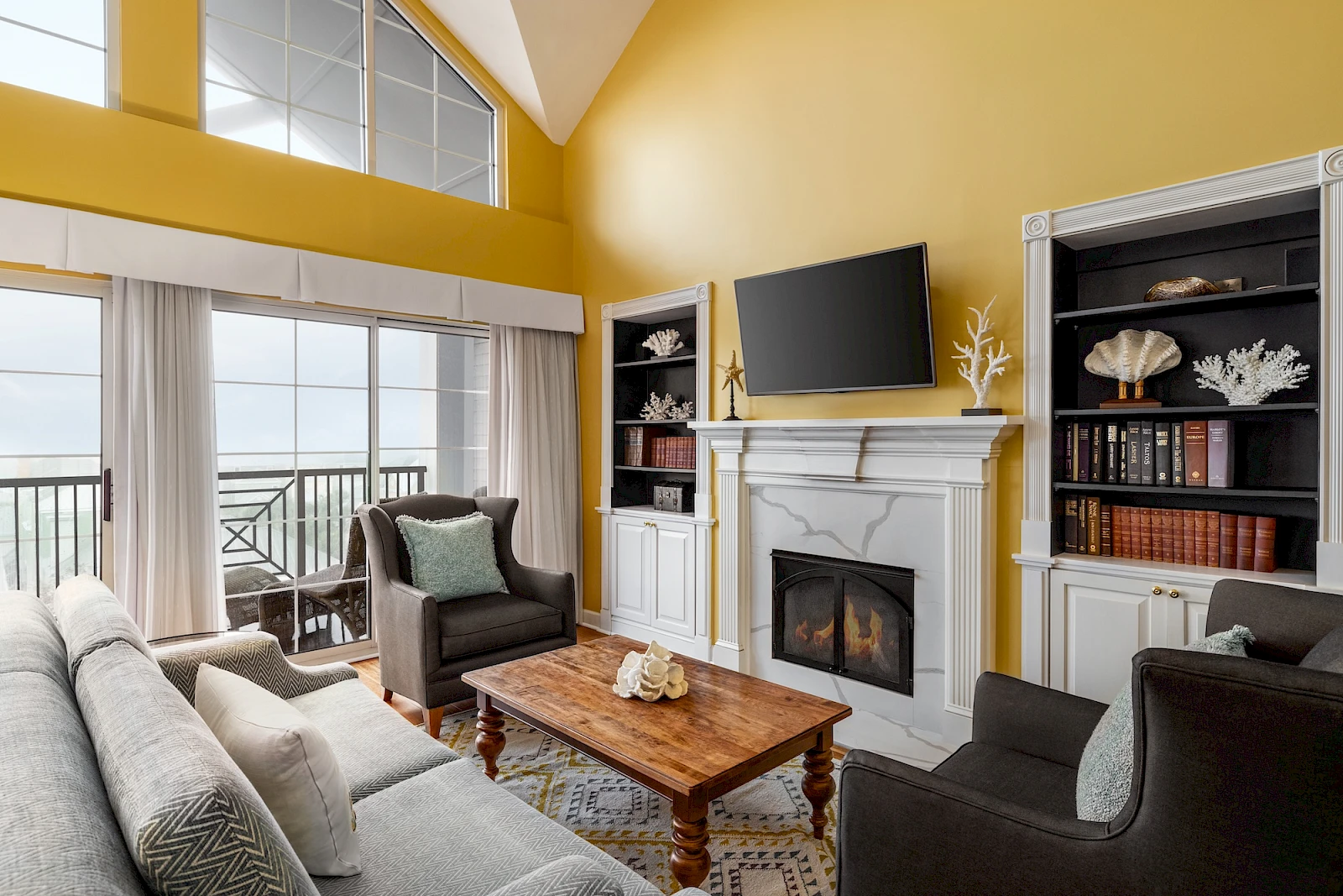 Cozy coastal living room with a bright yellow wall, a fireplace, built-in shelves, a flat-screen above the mantel, and seating facing a lake view balcony.