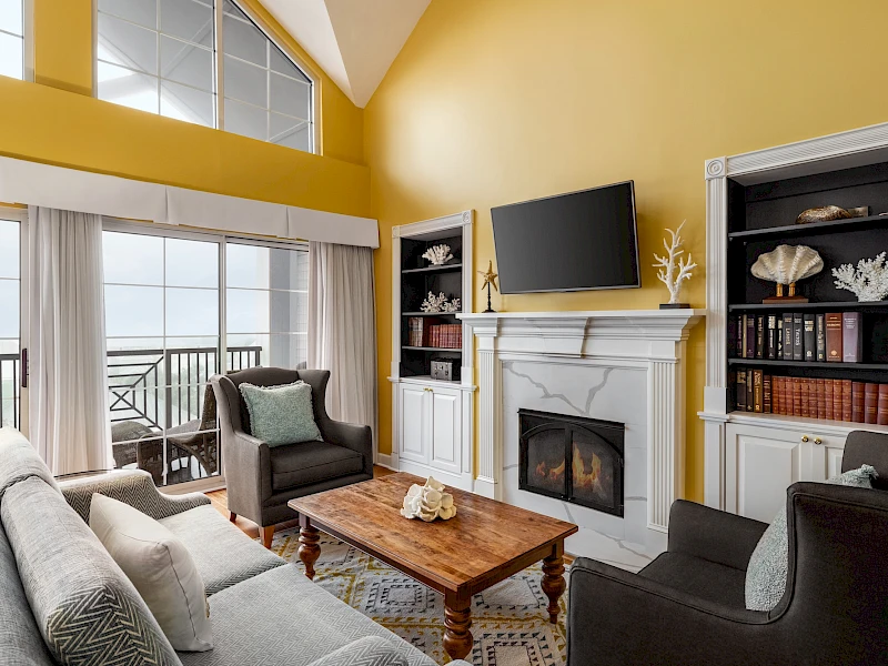 A bright coastal living room with a yellow vaulted ceiling, a white fireplace, built-in shelves, a wooden coffee table, and a view of the balcony oceanfront.