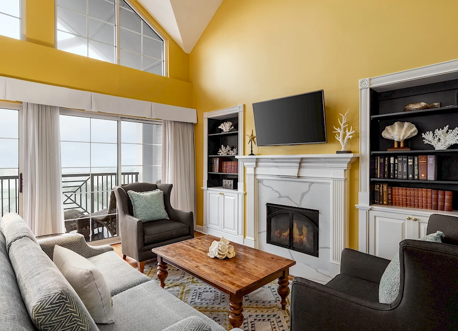 A bright coastal living room with a yellow vaulted ceiling, a white fireplace, built-in shelves, a wooden coffee table, and a view of the balcony oceanfront.