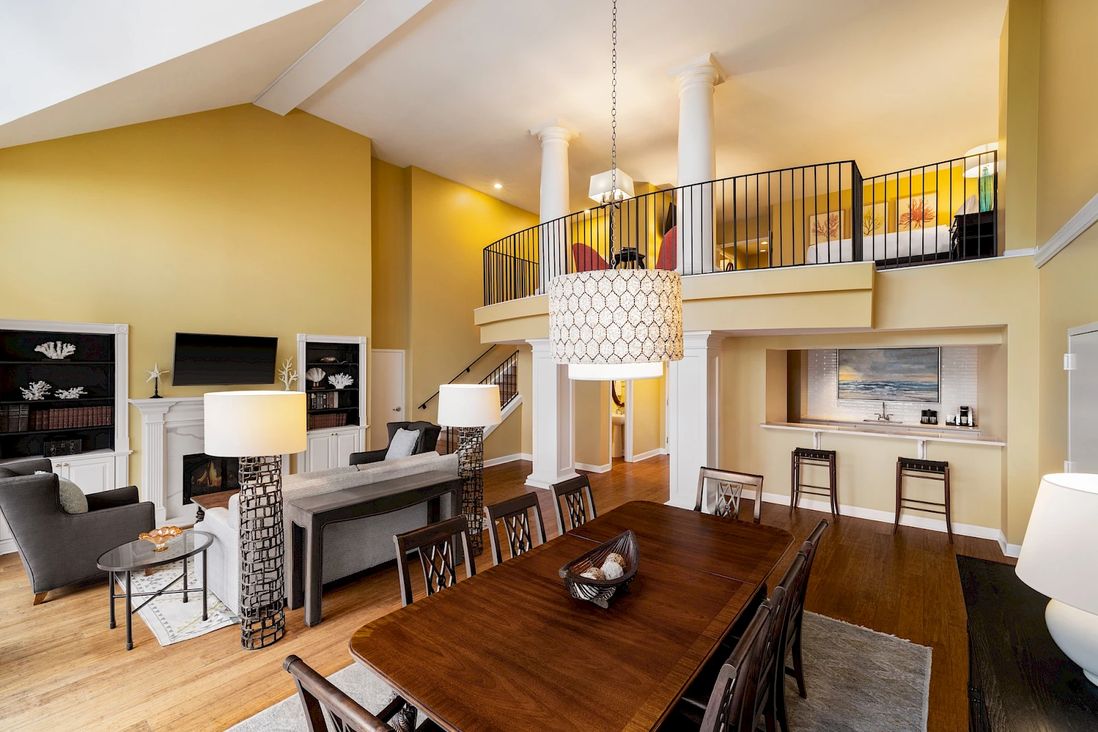 A bright, airy loft-style living space with a long wooden dining table, a piano bar area, yellow walls, and a second-floor balcony overlooking the room.