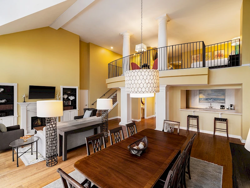 A bright, airy loft-style living space with a long wooden dining table, a piano bar area, yellow walls, and a second-floor balcony overlooking the room.
