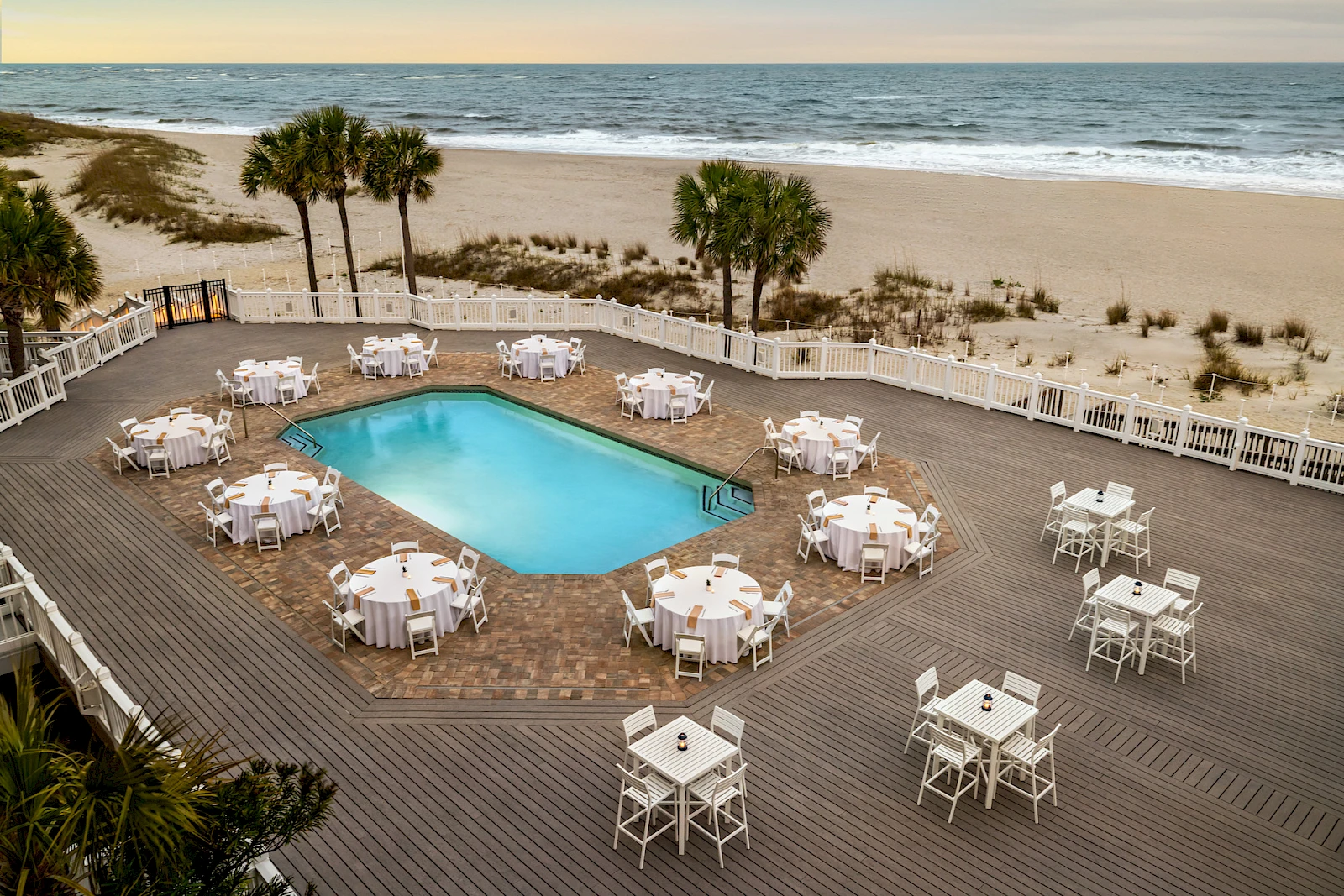 Beachside event setup around a pool: white round tables with chairs on a wooden deck, palm trees, sandy dunes, and the ocean in the background.