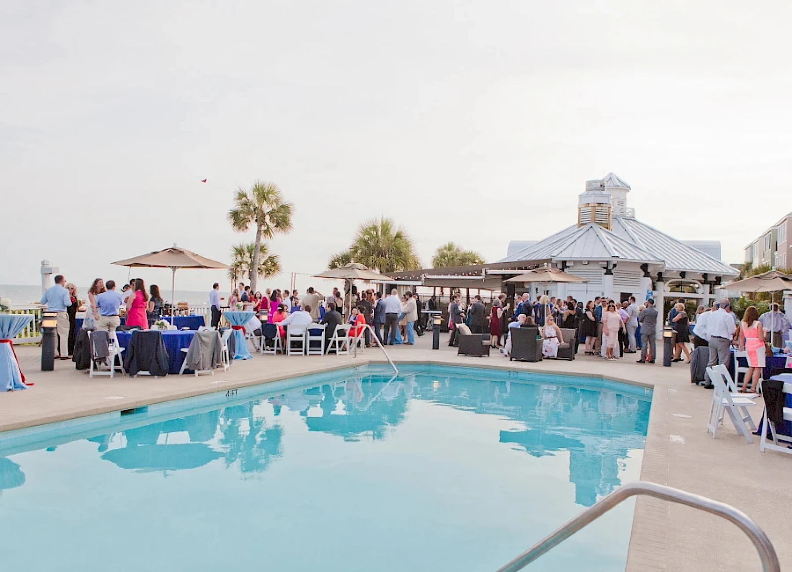A poolside party or wedding reception with guests mingling by tables and a gazebo, palm trees, and a bright, sunny outdoor setting.